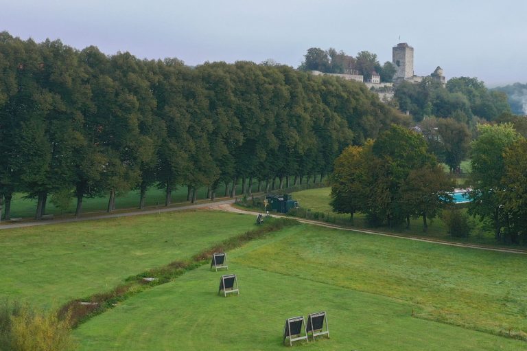 Landschaft mit Bäumen, einem Weg, einem Bach und einem historischen Gebäude im Hintergrund.