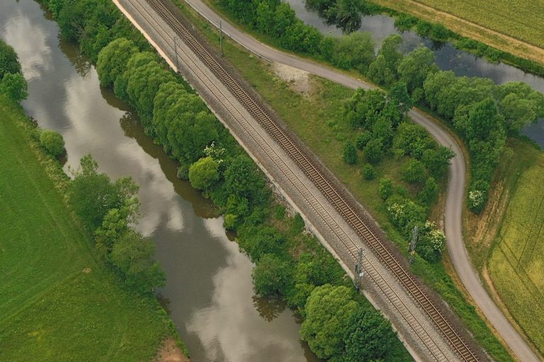 Eisenbahnschienen neben einem Fluss, umgeben von grünen Bäumen und Feldern.